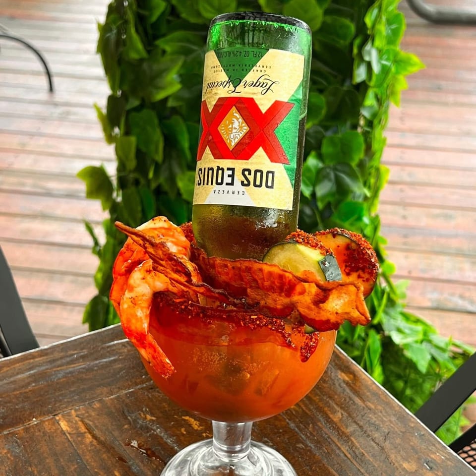 A Dos Equis beer bottle inverted into a michelada glass with shrimp and lime on a wooden table.