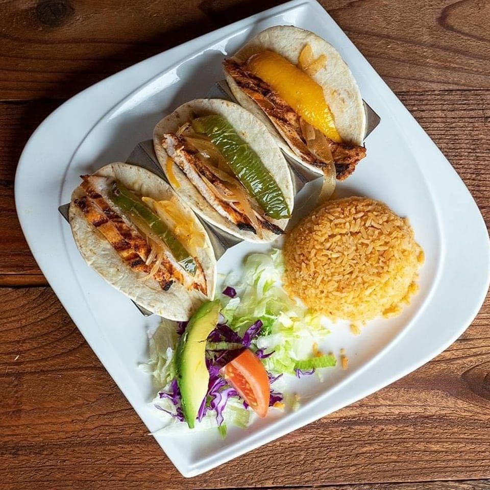 Three grilled chicken tacos with peppers and onions, served with rice, lettuce, avocado, and tomato.